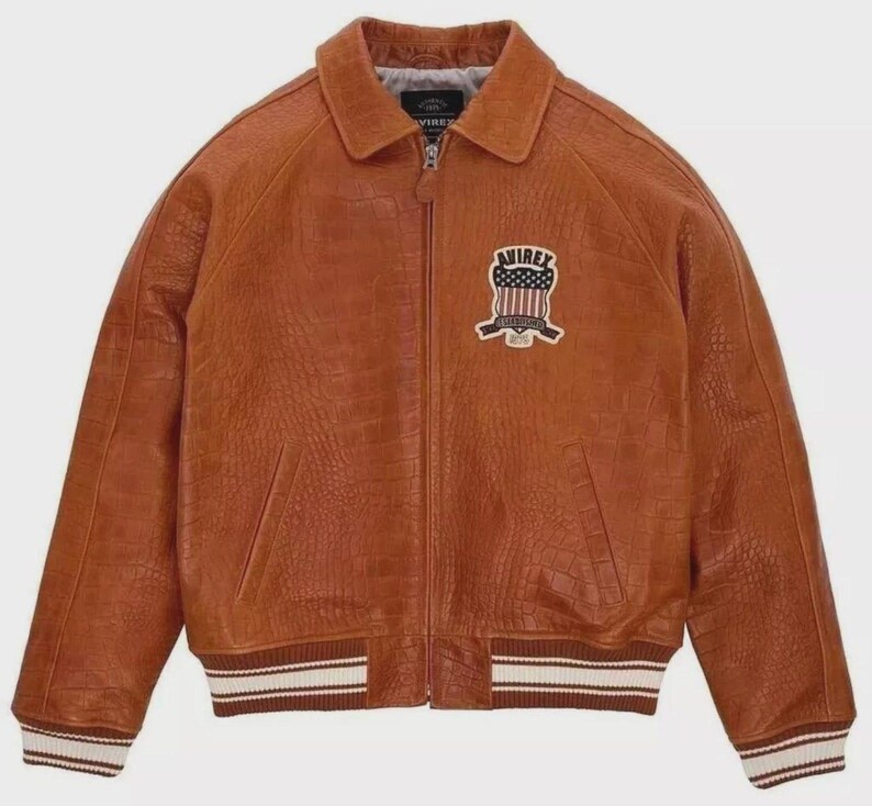 Puede incluir: Chaqueta de cuero marr&oacute;n con acabado texturizado, cuello y cierre de cremallera. La chaqueta tiene un parche en el pecho con un dise&ntilde;o de bandera americana y la palabra "AVIREX". Los pu&ntilde;os y el dobladillo tienen un dise&ntilde;o a rayas.