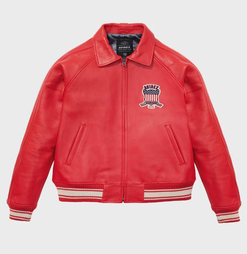May include: A vibrant red leather jacket with a classic bomber style. Features a collared neckline, a zipper closure, and a patch on the chest. The cuffs and hem have white and red striped detailing. The jacket has a logo patch that says "AVIREX".