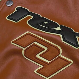 May include: Close-up of a brown leather jacket with large, stitched-on black and brown letter patches. The patches have a cream-colored border. A button with the text "GREEN USA VARSITY" is visible at the top.