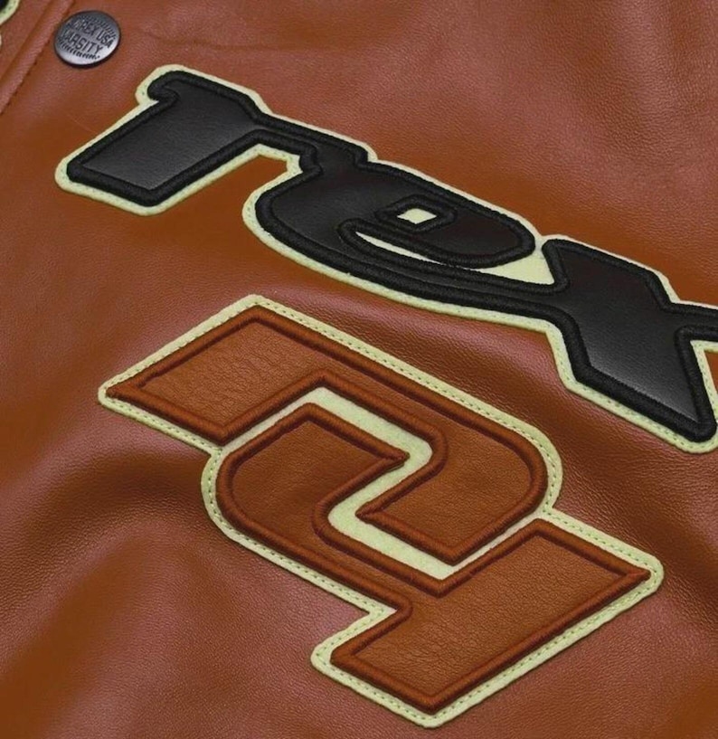May include: Close-up of a brown leather jacket with large, stitched-on black and brown letter patches. The patches have a cream-colored border. A button with the text "GREEN USA VARSITY" is visible at the top.