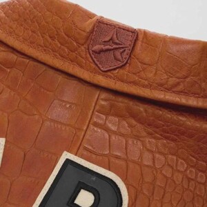 Puede incluir: Primer plano de una chaqueta de cuero marr&oacute;n con textura de cocodrilo. La chaqueta presenta un peque&ntilde;o emblema bordado y un parche de letras grande con borde blanco y negro. El cuero tiene un tono c&aacute;lido y rico.