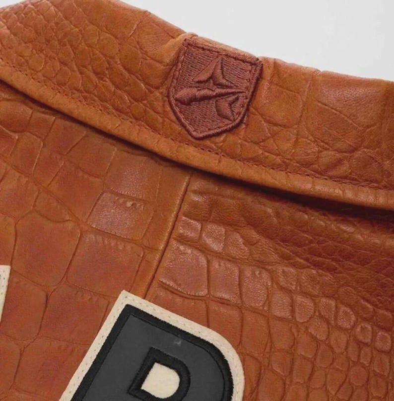 Puede incluir: Primer plano de una chaqueta de cuero marr&oacute;n con textura de cocodrilo. La chaqueta presenta un peque&ntilde;o emblema bordado y un parche de letras grande con borde blanco y negro. El cuero tiene un tono c&aacute;lido y rico.