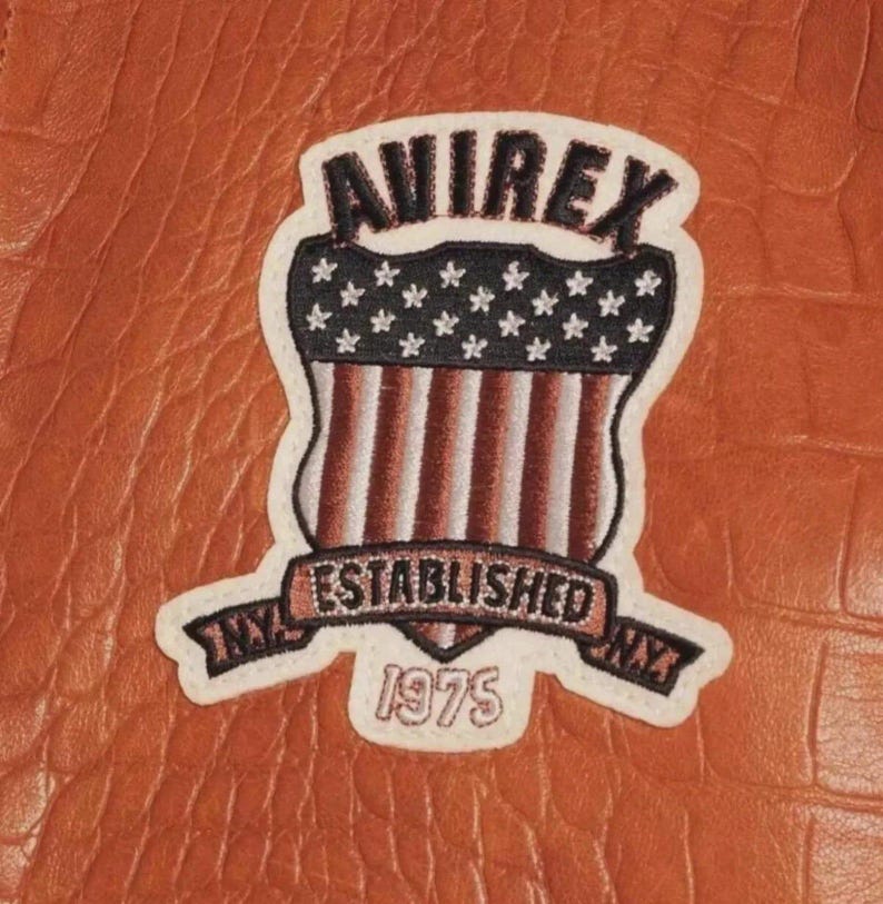 Puede incluir: Primer plano de un parche de cuero marr&oacute;n con el texto "AVIREX" en la parte superior. El parche presenta un dise&ntilde;o de escudo con la bandera estadounidense y las palabras "ESTABLISHED NY 1975" en una pancarta.