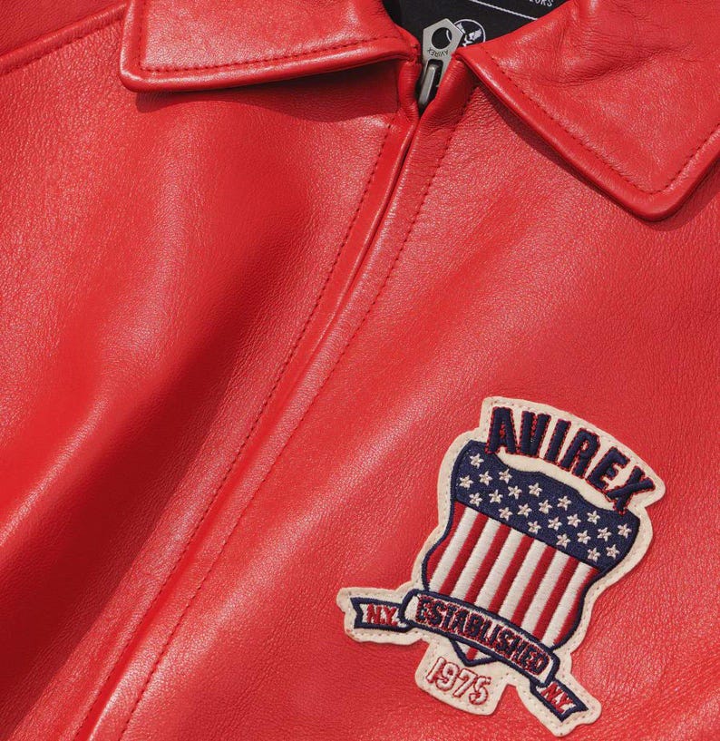 May include: Close-up of a bright red leather jacket with a collar and zipper. The jacket features a patch with the text "AVIREX" above a shield design with the American flag and the text "NY ESTABLISHED 1975".