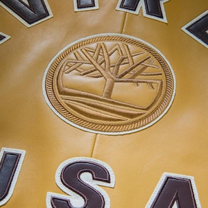 K&ouml;nnte beinhalten: Nahaufnahme einer senffarbenen Jacke mit braunen und wei&szlig;en gestickten Aufn&auml;hern. Der zentrale Aufn&auml;her zeigt ein Baumdesign in einem kreisf&ouml;rmigen Rahmen. Die Buchstaben "USA" sind sichtbar.