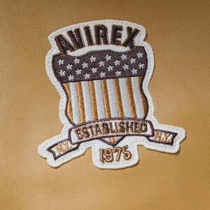 K&ouml;nnte beinhalten: Gestickter Aufn&auml;her mit dem Text "AVIREX" &uuml;ber einem Schilddesign mit Sternen und Streifen, mit "ESTABLISHED NY 1975" auf einem Banner. Der Aufn&auml;her ist wei&szlig; mit braunen, goldenen und wei&szlig;en N&auml;hten, vor einem beigen Hintergrund.