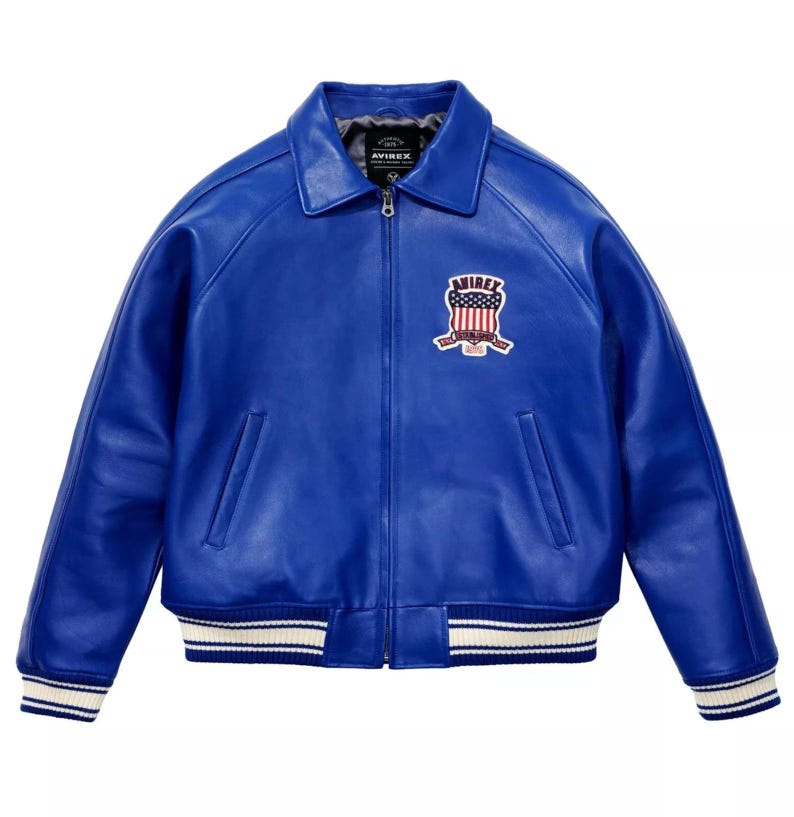 May include: A vibrant blue leather jacket with a classic collar and a zip-up front. The jacket features a white and blue striped trim on the cuffs and hem. An embroidered patch with the text "AVIREX" and a patriotic design is on the chest.