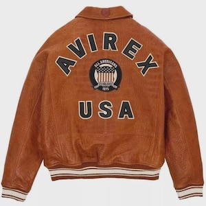 Puede incluir: Chaqueta bomber de cuero marr&oacute;n con la palabra "AVIREX" en letras negras y blancas en la espalda. Debajo, un parche con "ALL AMERICANS ESTABLISHED 1975" y un gr&aacute;fico de la bandera estadounidense. La palabra "USA" est&aacute; debajo. Cuello y pu&ntilde;os acanalados.