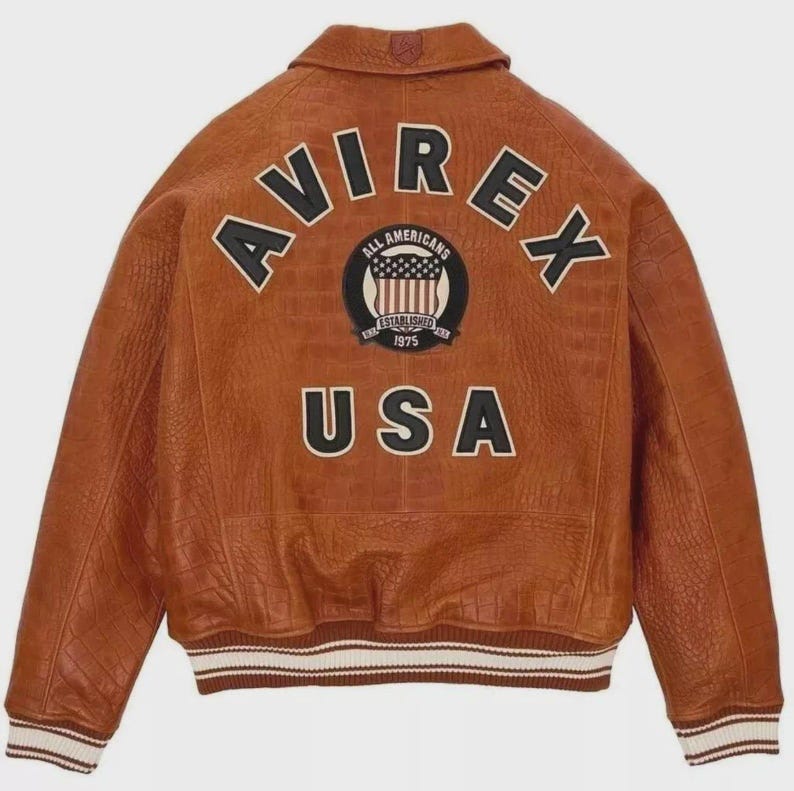Puede incluir: Chaqueta bomber de cuero marr&oacute;n con la palabra "AVIREX" en letras negras y blancas en la espalda. Debajo, un parche con "ALL AMERICANS ESTABLISHED 1975" y un gr&aacute;fico de la bandera estadounidense. La palabra "USA" est&aacute; debajo. Cuello y pu&ntilde;os acanalados.