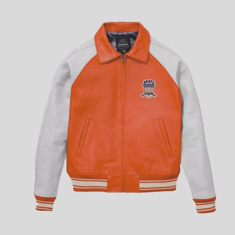 May include: Orange and white leather jacket with a zip-up front and a collared neckline. The jacket features white sleeves, a striped hem and cuffs, and a patch on the chest with the brand name. The jacket is a classic varsity style.