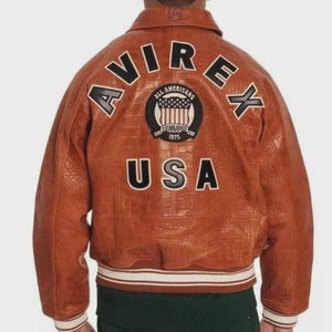 Puede incluir: Chaqueta de cuero marr&oacute;n con la palabra "AVIREX" en letras blancas en la espalda. Debajo, un parche dice "ALL AMERICANS ESTABLISHED 1975" y la palabra "USA". La chaqueta tiene un ribete acanalado blanco y crema.