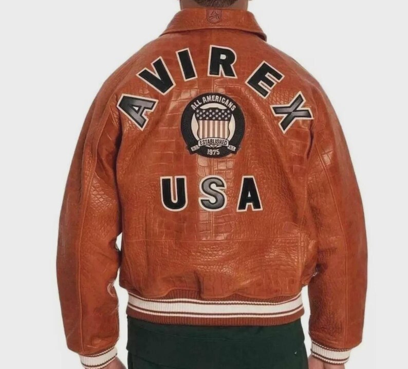 Puede incluir: Chaqueta de cuero marr&oacute;n con la palabra "AVIREX" en letras blancas en la espalda. Debajo, un parche dice "ALL AMERICANS ESTABLISHED 1975" y la palabra "USA". La chaqueta tiene un ribete acanalado blanco y crema.