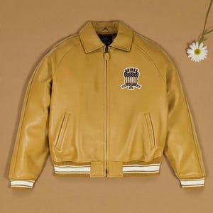 K&ouml;nnte beinhalten: Senfgelbe Lederjacke mit Rei&szlig;verschluss, Kragen und gerippten B&uuml;ndchen und Saum mit wei&szlig;en Streifen. Verf&uuml;gt &uuml;ber einen Aufn&auml;her auf der Brust mit dem Text "AVIREX" und einem Schilddesign. Ein klassischer Bomberjacken-Stil.