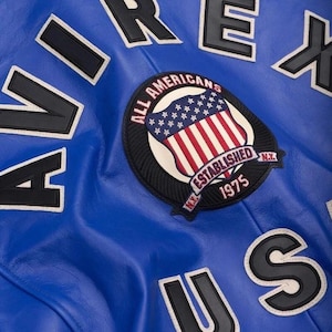 May include: Close-up of a blue leather jacket with white and black lettering spelling out "AIREX US". A circular patch features the American flag and the text "ALL AMERICANS ESTABLISHED 1975 N.Y."