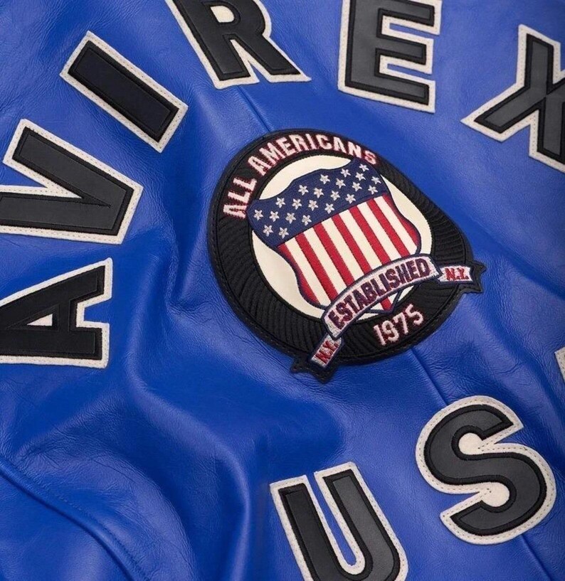 May include: Close-up of a blue leather jacket with white and black lettering spelling out "AIREX US". A circular patch features the American flag and the text "ALL AMERICANS ESTABLISHED 1975 N.Y."