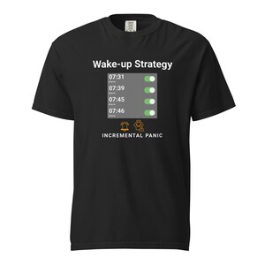 May include: Black t-shirt featuring the words "Wake-up Strategy" and a graphic of a phone screen showing alarm times. The screen displays four alarms set for 07:31, 07:39, 07:45, and 07:46. Below is the text "INCREMENTAL PANIC".