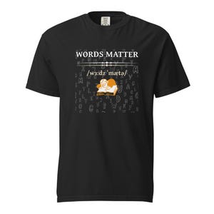 May include: Black t-shirt with the words "WORDS MATTER" in white, above a phonetic transcription. Below the text is a graphic of a person reading a book. The background is filled with scattered letters.