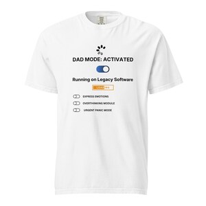 Op de afbeelding: Wit t-shirt met de tekst "DAD MODE: ACTIVATED" en een afbeelding van een laadbalk. Het shirt bevat ook de zinnen "Running on Legacy Software", "Express Emotions", "Overthinking Module" en "Urgent Panic Mode".