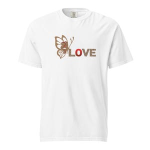 Può includere: T-shirt bianca con un disegno a farfalla e la scritta "LOVE". La farfalla è marrone con dettagli rossi, e le lettere "L", "O", "V" e "E" sono in stampa leopardata e rosse.