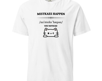 Könnte beinhalten: Weißes T-Shirt mit dem Text "MISTKAES HAPPEN" über der phonetischen Schreibweise "/mi'steiks 'hæpən/" und "YOU NOTICED". Darunter befindet sich ein Cartoon-Katzen-Gesicht mit einem Augenzwinkern.
