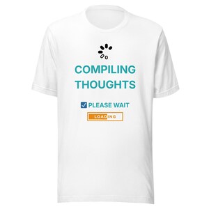Può includere: T-shirt bianca con la scritta "COMPILING THOUGHTS" in turchese, un'icona di caricamento e le frasi "PLEASE WAIT" e "LOADING". Il design è centrato sulla parte anteriore della maglietta.