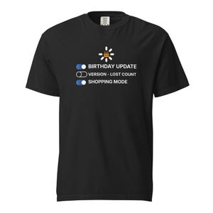 May include: Black t-shirt with a graphic design featuring a birthday cake icon and the text "BIRTHDAY UPDATE", "VERSION - LOST COUNT", and "SHOPPING MODE". The design is in white and light blue.