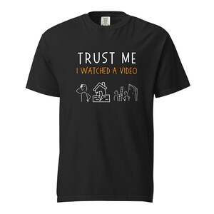 May include: Black t-shirt with the text "TRUST ME I WATCHED A VIDEO" in white and orange. The shirt features a humorous stick figure illustration of a person, a house, and people on a ladder. A casual, comfortable tee.