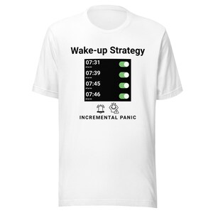 May include: White t-shirt featuring the words "Wake-up Strategy" and a graphic of a phone screen with multiple alarms set for different times. The phrase "Incremental Panic" is printed below the graphic.