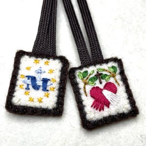 May include: Two embroidered religious pendants with brown cords. One features a blue "M" with a cross and gold stars, the other a heart, cross, and leaves. Both have a white background and brown borders.