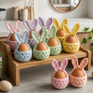 Puede incluir: Una colección de coloridos portahuevos de conejito de ganchillo en tonos pastel de rosa, azul, verde, amarillo y morado. Cada soporte contiene un huevo marrón y presenta orejas de conejo y un lazo. Se exhiben en una bandeja de madera.