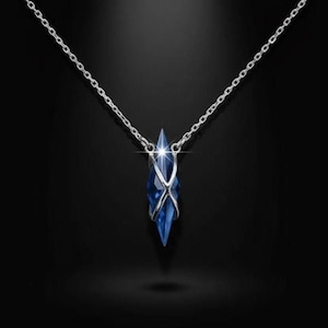 Può includere: Una collana a catena color argento con un ciondolo in cristallo blu intenso sfaccettato, racchiuso in una delicata gabbia di metallo argentato incrociata. Il ciondolo ha una forma a diamante allungata e appuntita con un riflesso luminoso.