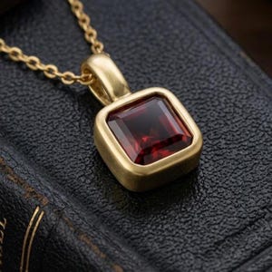 May include: A gold pendant necklace featuring a square-cut deep red garnet gemstone set in a brushed gold frame. The pendant is attached to a delicate gold chain, showcasing a smooth, matte finish on the metal setting surrounding the stone.
