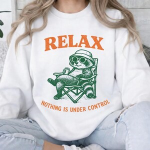 Puede incluir: Sudadera blanca con un gráfico naranja "RELAX" y una ilustración verde de un mapache con gafas de sol y sombrero, sentado en una silla, con el texto "NOTHING IS UNDER CONTROL".