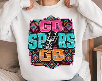 May include: White sweatshirt with a colorful graphic that says "GO SPURS GO". The graphic features the team's logo and a geometric design in pink, teal, orange, and black. The sweatshirt is a crew neck style.
