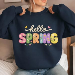 May include: Navy blue sweatshirt with the words "hello SPRING" in colorful, embroidered letters. The word "hello" is in a cursive font, and the word "SPRING" is in a block letter style with floral accents. The sweatshirt is a crew neck style.