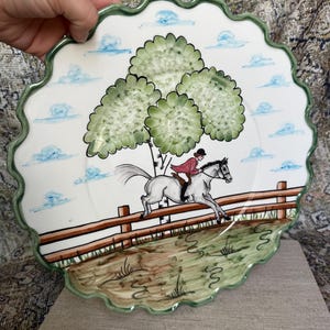 May include: White ceramic decorative plate with a scalloped green edge. The plate features a hand-painted scene of a person riding a white horse jumping over a fence, with green trees and blue clouds in the background.