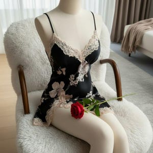 May include: Black satin slip dress with a floral pattern and lace trim. The dress features spaghetti straps and a V-neckline. A red rose rests on the mannequin's leg. The dress is displayed on a mannequin sitting in a fluffy white chair.