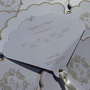 May include: White wedding fan with a scalloped edge and gold trim. The fan displays the wedding itinerary, including ceremony, meal, and reception times. The text includes the names Harvey & Lila and the date.