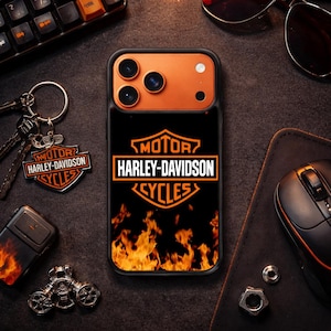May include: Black phone case featuring the Harley-Davidson Cycles logo in orange and white, with a flame design. The case is surrounded by a keyboard, sunglasses, a keychain, a lighter, and a computer mouse.