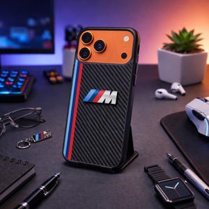 May include: Black iPhone case with an orange top and carbon fiber texture. Features a vertical red, blue, and white stripe and a BMW M logo. The case is displayed on a black stand, with a keychain and other accessories in the background.