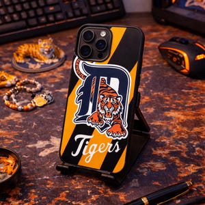 Puede incluir: Funda de teléfono negra con un diseño a rayas amarillas y negras. La funda presenta un logotipo de los Tigres de Detroit con un gráfico de tigre y la palabra "Tigers" en blanco. La funda está sobre un soporte negro.