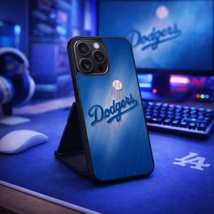 May include: A blue phone case featuring the Los Angeles Dodgers logo with a baseball and the word "Dodgers" in white script. The case is displayed on a black stand, with a computer and accessories in the background.