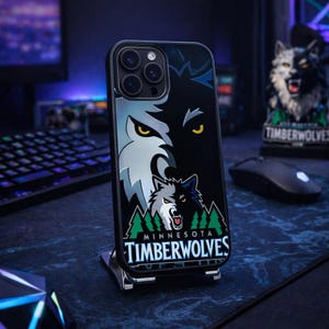 Puede incluir: Funda de teléfono negra con un diseño de lobo y el texto "MINNESOTA TIMBERWOLVES". La funda tiene un borde negro y un soporte plateado. El diseño incluye la cara de un lobo, árboles y el logotipo del equipo.
