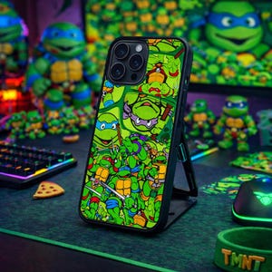 May include: A black smartphone case featuring a collage of green Teenage Mutant Ninja Turtles characters. The case is displayed on a stand, with a green and black gaming mouse, keyboard, and figurines in the background. A green wristband with "TMNT" is also visible.