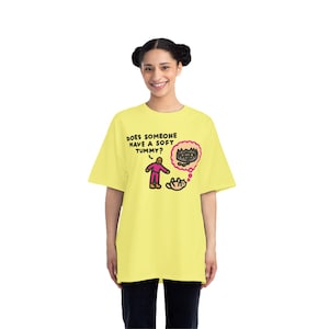 May include: A light yellow t-shirt with the text "DOES SOMEONE HAVE A SOFT TUMMY?" and a cartoon illustration. The illustration features a pink figure, a thought bubble with a brown object, and a white cat.
