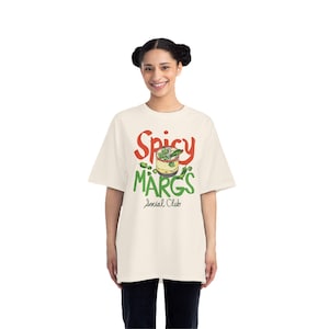 Puede incluir: Camiseta color crema con las palabras "Spicy Margs Social Club" en rojo y verde. La camiseta presenta un gráfico de una margarita con jalapeños. La modelo lleva pantalones negros.