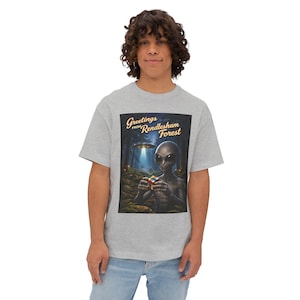 May include: A light gray t-shirt featuring a graphic of an alien holding a Rubik's Cube in a forest setting with a UFO. The text "Greetings from Rendlesham Forest" is arched above the image.