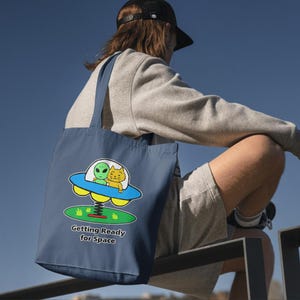 May include: Navy blue tote bag featuring a cartoon illustration of an alien and a cat in a UFO. The UFO is on a spring, with the text "Getting Ready for Space" below. The bag has two blue handles.