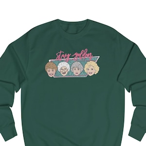 May include: Dark green sweatshirt featuring a graphic of four women's faces with different hairstyles. Above the faces is the text "Stay Golden" in pink script within a teal and white design.