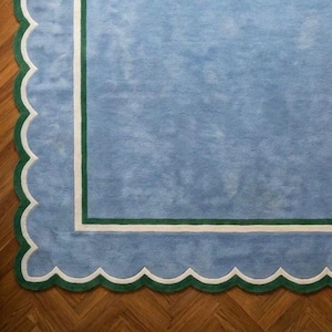 Puede incluir: Una alfombra azul claro con un borde festoneado blanco y verde. La alfombra tiene forma rectangular con un borde blanco y verde. La alfombra está colocada sobre un suelo de madera.